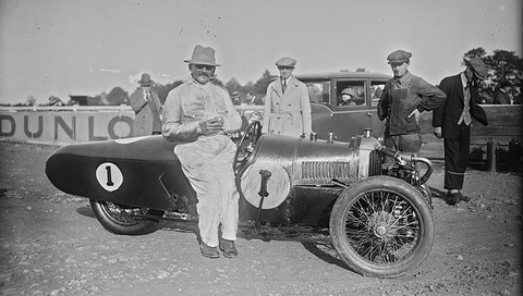A heavy man in white overalls and a hat leans on a long-tailed racing three-wheeler numbered 1, men and a Dunlop banner behind. Opens a larger view