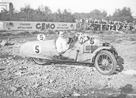 A smiling driver in a flower-garlanded three-wheeled racing car numbered 5 on stony ground, a crowd behind a fence and a Gémo hoarding. Opens a larger view