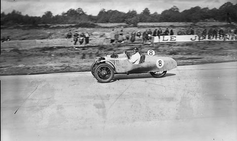 A three-wheeled racing car numbered 8 on a concrete track, a few spectators and a newspaper banner behind. Opens a larger view
