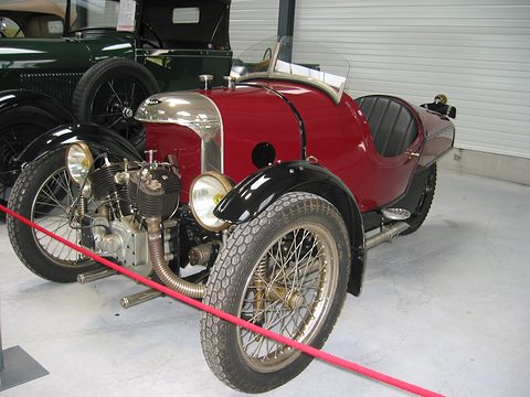 A red three-wheeler with a polished nose cowl, two headlamps and an air-cooled V-twin ahead of the front wheels, behind a red rope. Opens a larger view