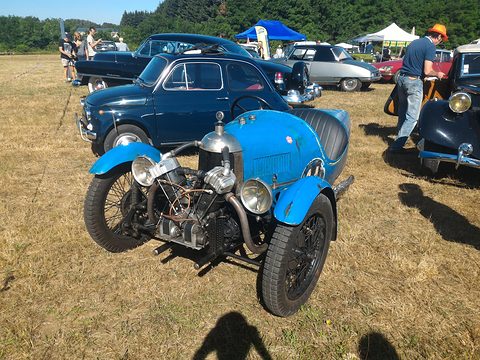 A blue three-wheeler with an exposed V-twin on dry grass, older saloons and people behind, a shadow in front. Opens a larger view