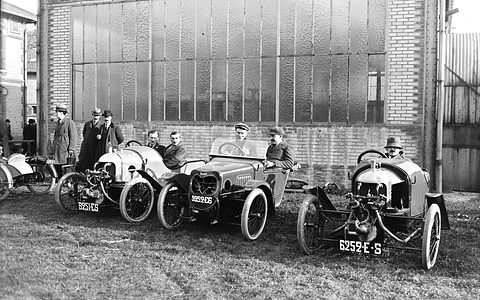 Three small three-wheelers parked in a row on grass in front of a tall glazed hangar, drivers and onlookers in caps around them. Opens a larger view