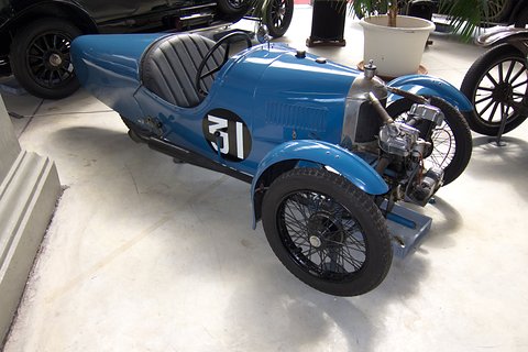 Side view of a blue racing three-wheeler with a black pleated seat, louvred bonnet and exposed engine. Opens a larger view