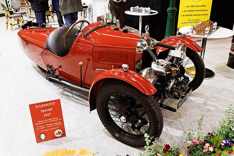 A red three-wheeler with a black seat and exposed engine, a red placard beside it and café chairs behind. Opens a larger view
