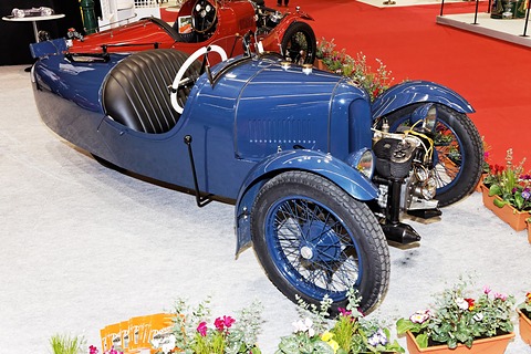 Side view of a dark blue three-wheeler with a pleated black seat and exposed engine, flowers in the foreground. Opens a larger view