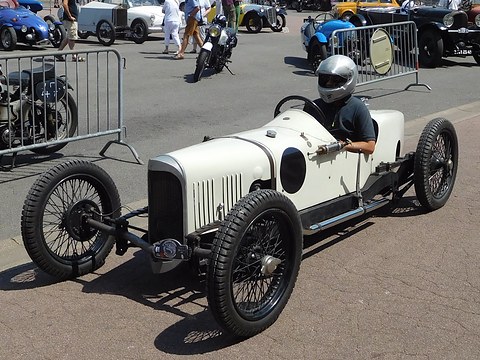 A white three-wheeler with a louvred bonnet and black wheels, a helmeted driver aboard, barriers and motorcycles behind. Opens a larger view