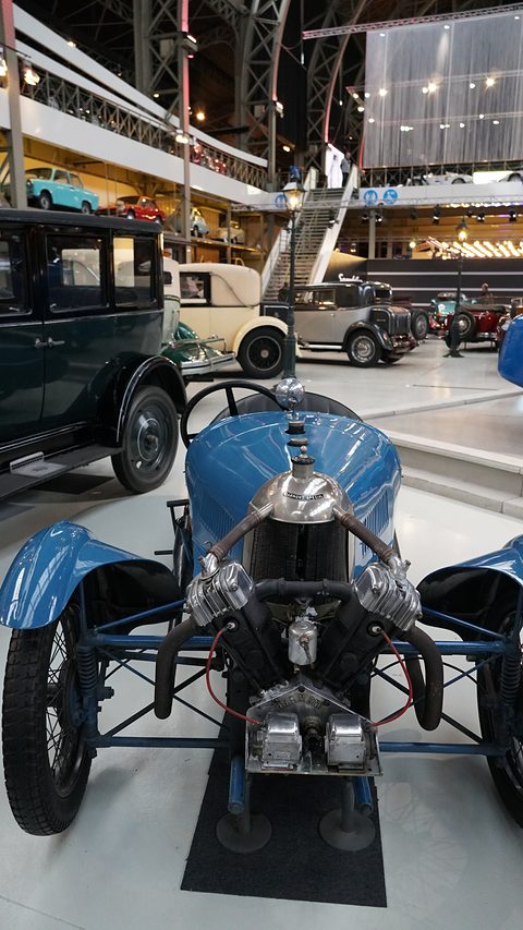 Front view from above of a blue three-wheeler with a V-twin, in a hall with a high iron roof and other cars. Opens a larger view