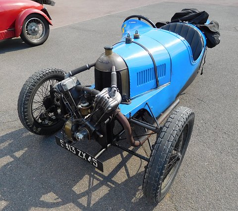 A blue three-wheeler with a strapped bonnet, exposed V-twin and number plate, a red car behind. Opens a larger view