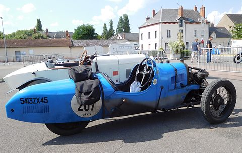 Side view of a blue racing three-wheeler with a bag strapped behind the seat, a white sports car and a town square behind.