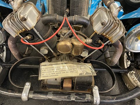 Close view of a finned V-twin engine with a brass carburettor, red plug leads and a printed card below. Opens a larger view