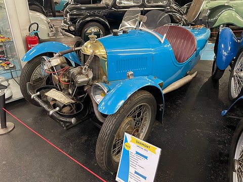 A blue three-wheeler with brass radiator cowl, maroon seat and exposed engine, a yellow and white label in front. Opens a larger view