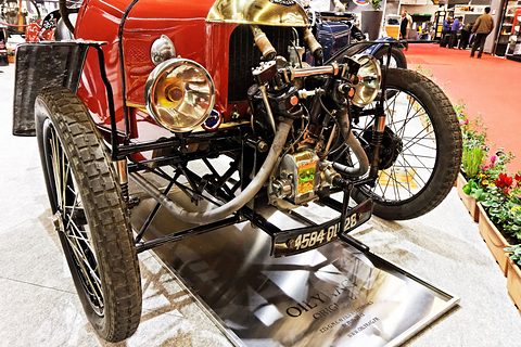 Close front view of a red three-wheeler: brass headlamps, an exposed V-twin engine and coil-sprung front wheels.