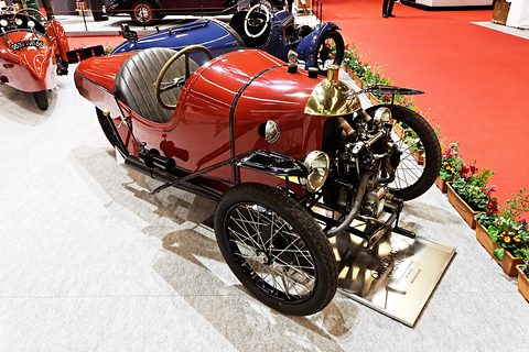 Looking down on a red three-wheeler with a brass cowl, exposed engine and wire wheels beside a bed of flowers. Opens a larger view