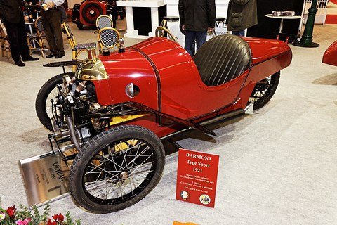 A red three-wheeler with a black buttoned seat and brass radiator cowl, a red placard and a steel sign before it, visitors behind. Opens a larger view