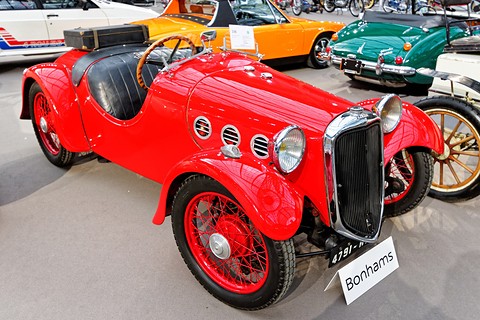 A red four-wheeled sports car with porthole vents, a chrome grille and red wire wheels, a sale sign in front. Opens a larger view