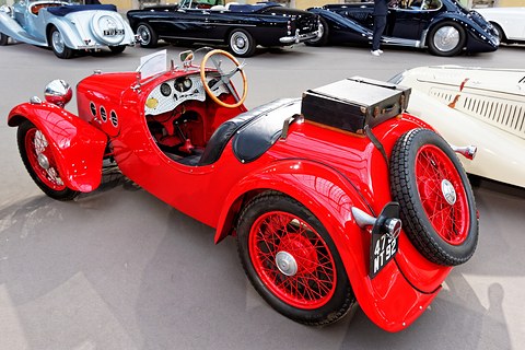 Rear quarter view of a red sports car with a spare wire wheel and a black trunk on the tail. Opens a larger view