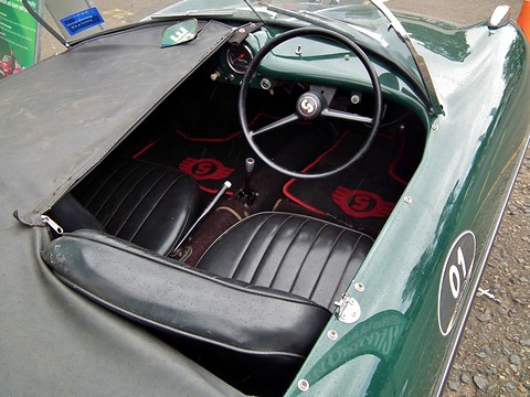 Looking down into a green roadster: a thin steering wheel, a small dashboard and black pleated seats. Opens a larger view