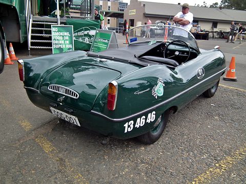 Rear three-quarter of a dark green roadster with a number on its flank, a trailer behind. Opens a larger view