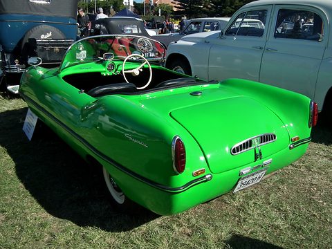 Rear three-quarter of a bright green open roadster on grass among other old cars. Opens a larger view