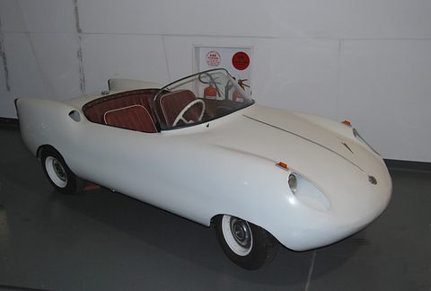 White open roadster with red seats on a grey museum floor against a white wall. Opens a larger view