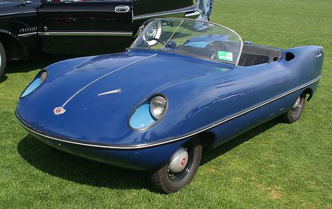 A low blue two-seat roadster with recessed round headlamps, a wrap-round screen and no doors, on a lawn. Opens a larger view