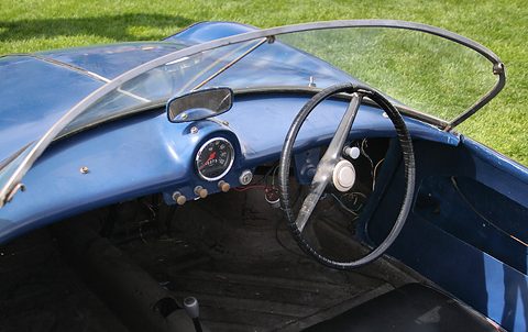 Looking into a blue roadster: a thin black steering wheel, one round speedometer and three knobs on a curved dash. Opens a larger view