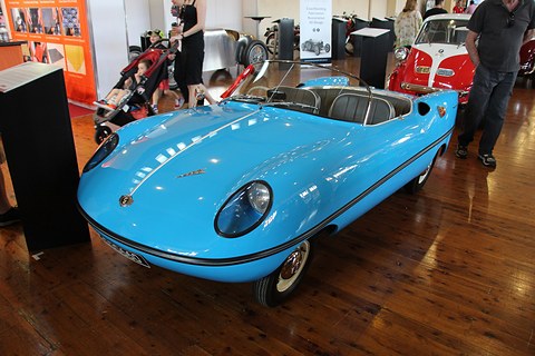 Light blue open roadster on a polished wooden floor in a showroom with posters behind. Opens a larger view