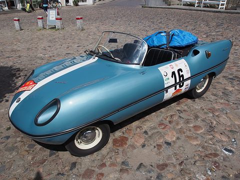 Light blue open roadster with a white stripe and number 16, luggage in its cockpit, on cobblestones.