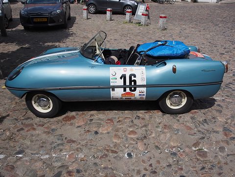 Side view of a light blue doorless roadster with a number 16 roundel on its flank, on cobbles. Opens a larger view