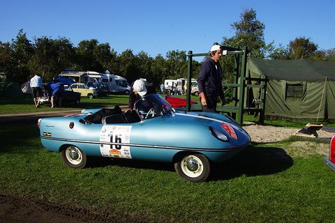 Light blue roadster with a number 16 roundel on grass, a man and tents behind. Opens a larger view