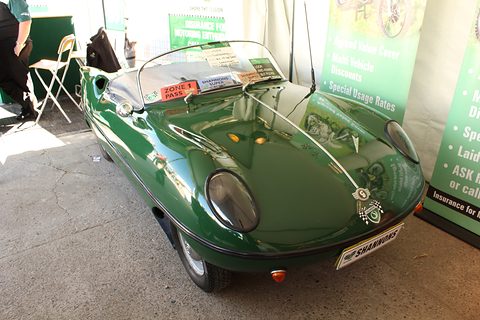 Dark green open roadster on a show stand in front of a green Shannons banner. Opens a larger view