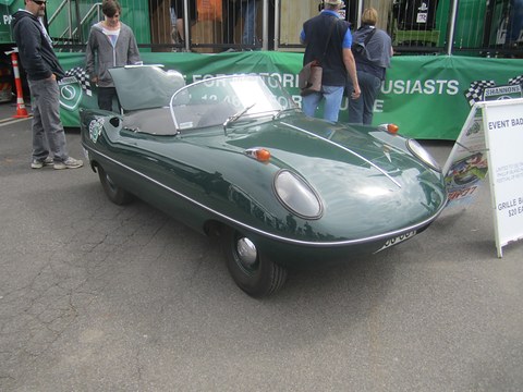 Dark green open roadster with an event board beside it and visitors in front of a green banner. Opens a larger view