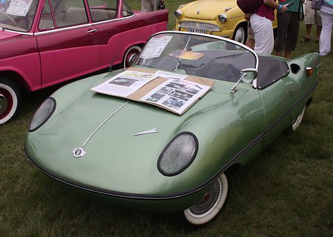 Pale green open roadster on grass with a folder of photographs on the bonnet, a pink car behind. Opens a larger view