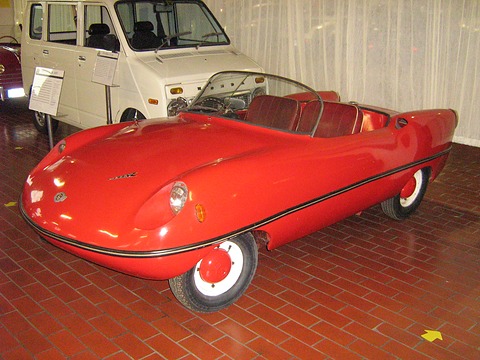Red doorless two-seat roadster with a wraparound windscreen and white wheels on a tiled floor. Opens a larger view
