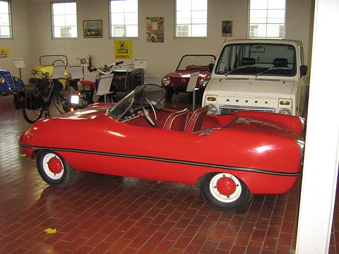 Side view of a red doorless roadster on a brick-red tiled floor, a white van behind.