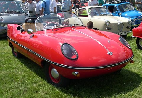 Red open roadster on grass with other small cars and visitors behind. Opens a larger view