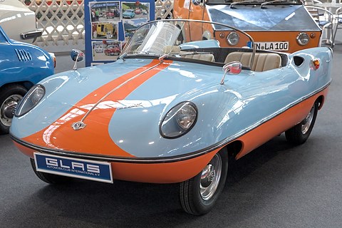 Pale blue roadster with an orange stripe and a GLAS plate on a show stand. Opens a larger view