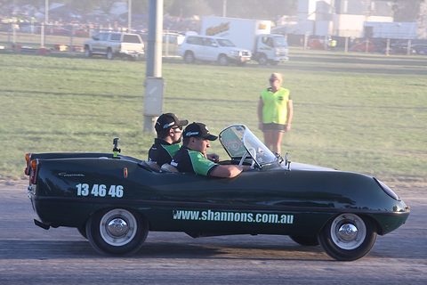 A low dark green doorless roadster with two men in caps aboard, driving past a hi-vis marshal on a dirt track. Opens a larger view