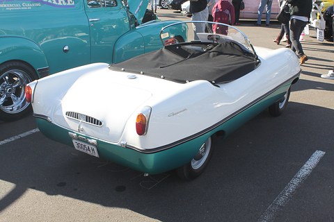 Rear three-quarter of a white and teal roadster with a black tonneau, on tarmac. Opens a larger view