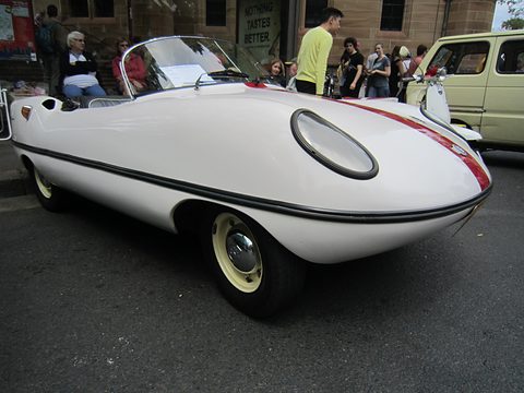 White open roadster with a red stripe from the front left, a crowd of visitors behind. Opens a larger view