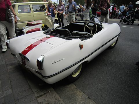 Rear three-quarter of a white open roadster with a red stripe, people walking past behind. Opens a larger view