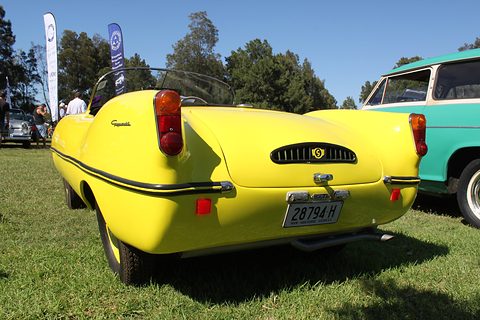 Rear view of a yellow open roadster with a slotted engine grille, parked on grass under a blue sky. Opens a larger view