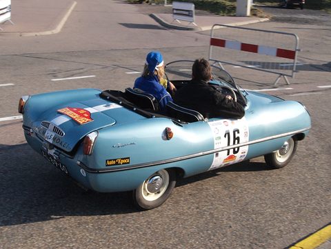 Light blue open roadster with two occupants and a number 16 roundel, driving past a barrier. Opens a larger view