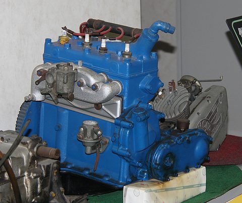 A blue four-cylinder side-valve engine with its gearbox on a museum display stand. Opens a larger view