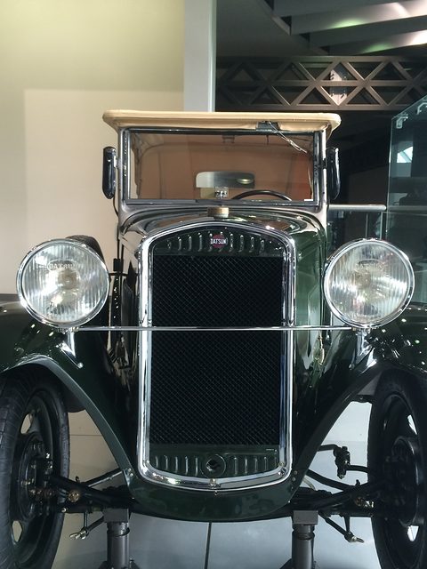 Head-on view of a dark green car’s narrow mesh radiator between two large round headlamps and curved black wings. Opens a larger view