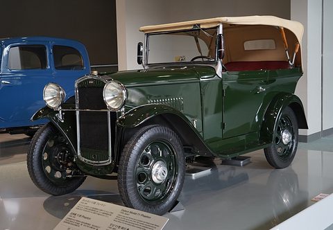 A dark green open tourer with a tan hood and red seats seen from the front three-quarter, a blue car behind it. Opens a larger view