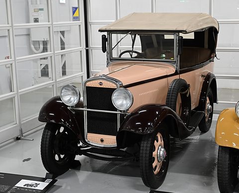 A small pale pink open tourer with maroon wings and a tan hood stands in a white garage hall.