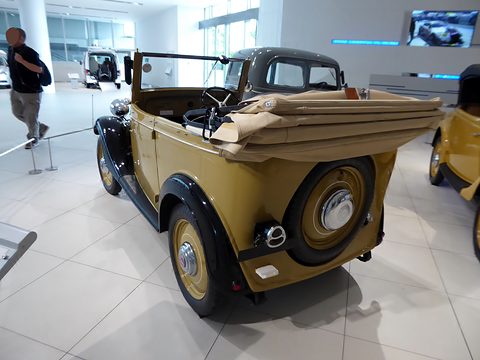 The rear quarter of a yellow open tourer with black wings, a folded tan hood and a spare wheel. Opens a larger view