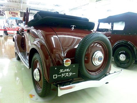 The rear of a maroon roadster with a black hood, a spare wheel and a Japanese name board on its bumper. Opens a larger view