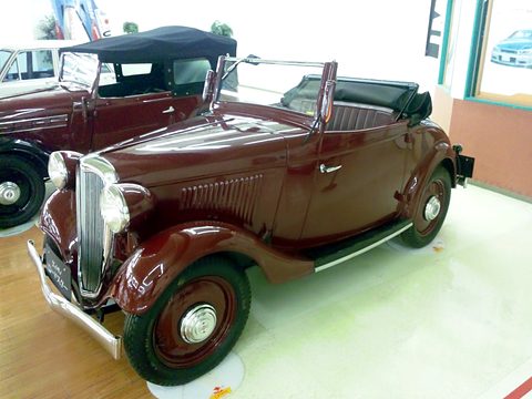A maroon two-seat roadster with its black hood folded, a chrome grille and white-wall tyres on a pale floor. Opens a larger view
